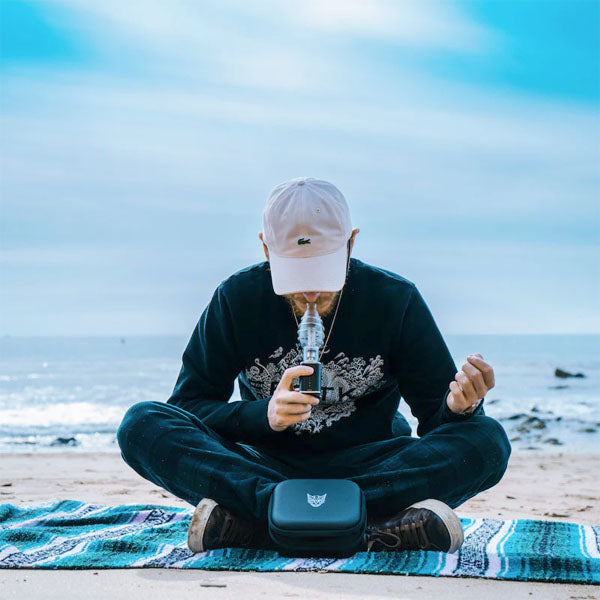 Linx Apollo Electric Dab Rig and E Nail in Portable Dab Rig mode with Linx Bubbler in use by a man sitting on a blanket on the beach with the ocean in the background